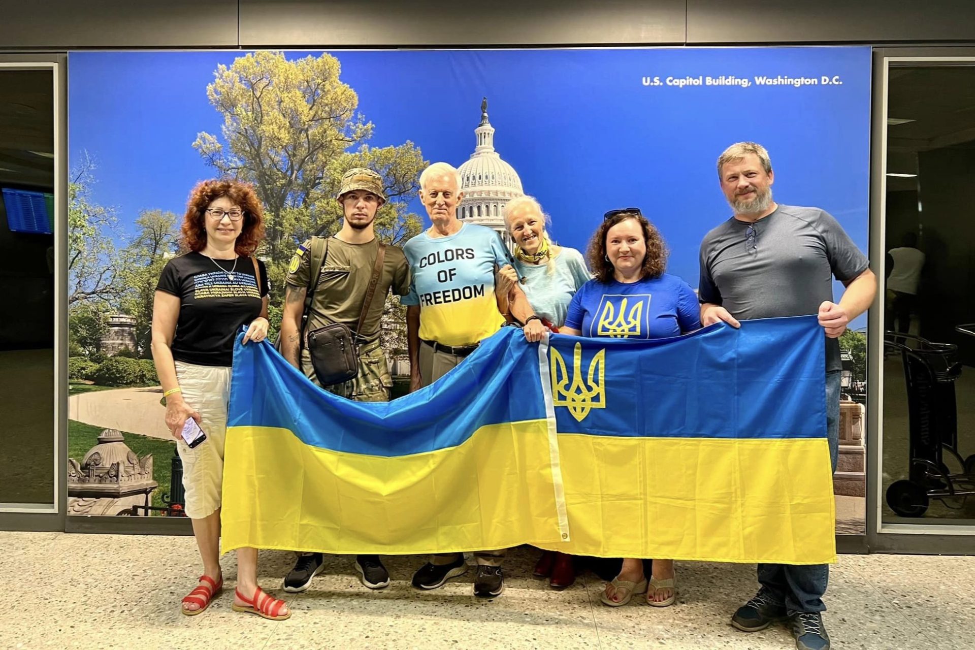 Ukrainian amputee veteran and hero, Heorhii Roshka surrounded by a group of 5 volunteers while Welcoming him in Washington, DC. All people in the photo are holding a big Ukrainian flag in front of them . On the background there's a photo of the Capitol in Washington