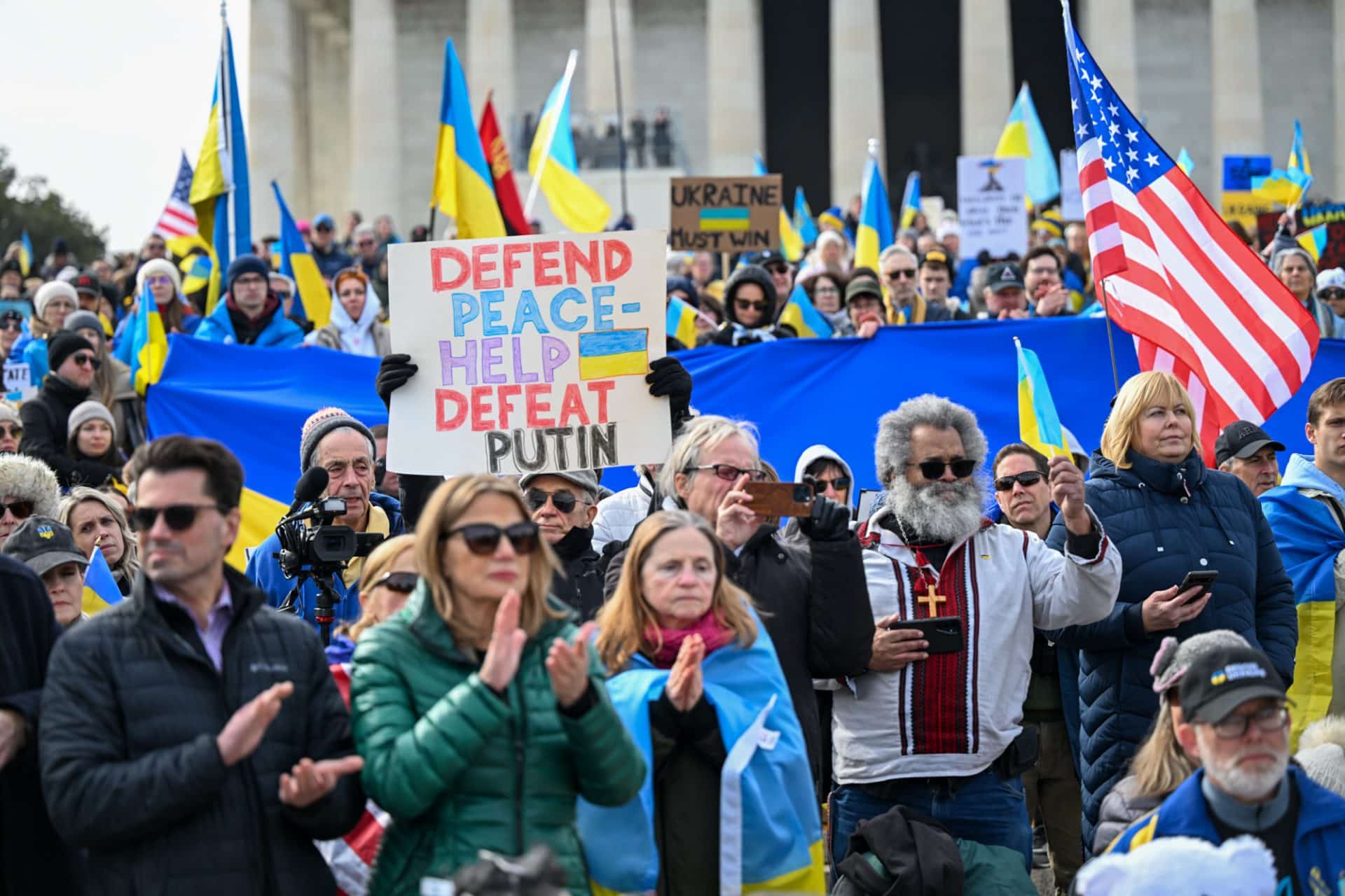 Defend Peace—Help Defeat Putin banner in a crowd of people in front of the Capitol. People handle a lot of American and Ukrainian flags.