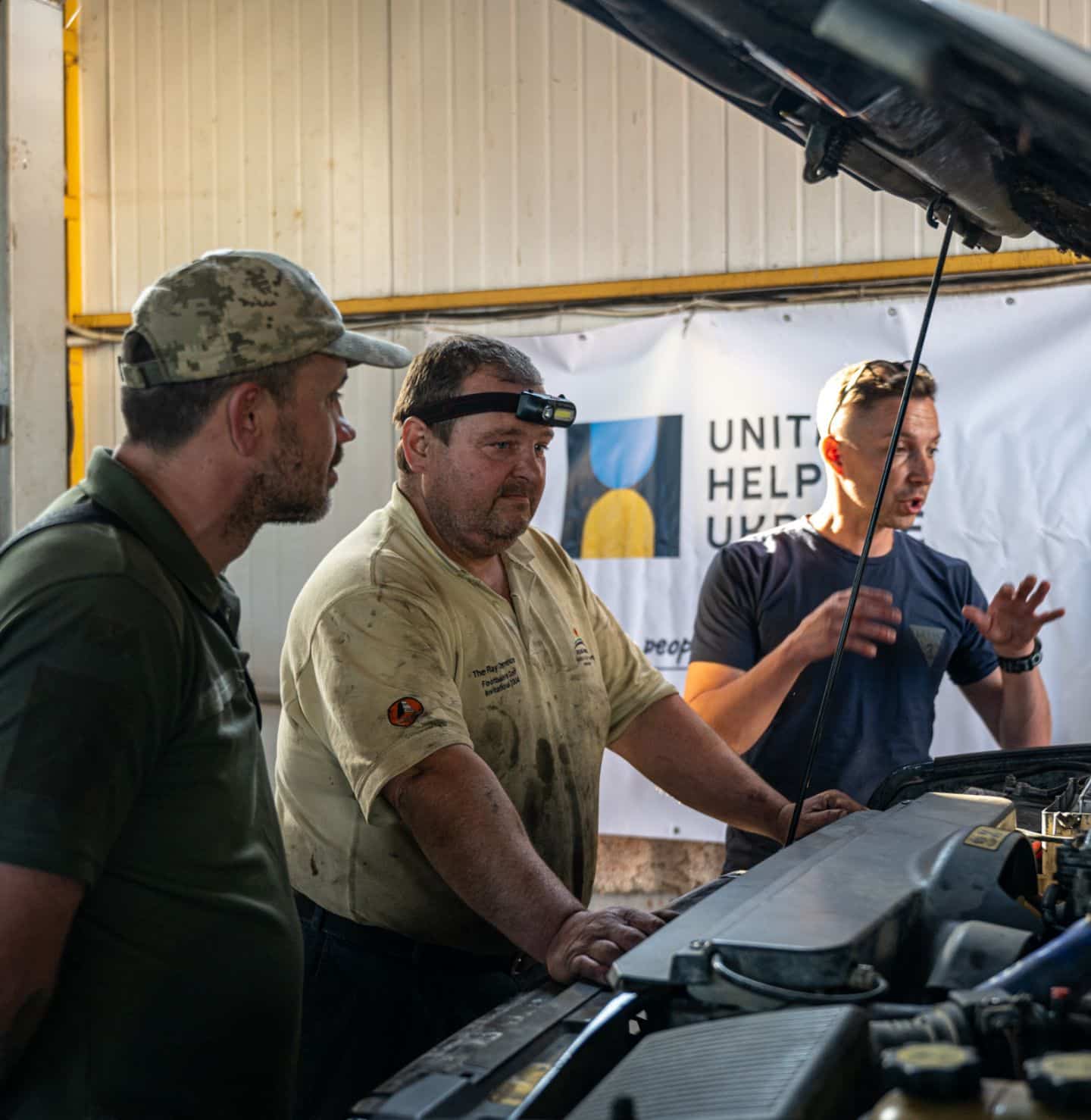 Car mechanics are inspecting a car in their garage to repair it and send it to the front lines. A banner with the Civilians HELP logo is in the background.